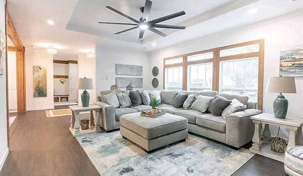 Elegant living room with a large gray sectional sofa and matching ottoman on a patterned rug. Sunlit windows, modern art, and a ceiling fan add charm.