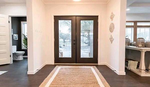 Spacious entryway with dark double doors, a woven rug on dark wood floors, and white walls adorned with decorative art. Calm and welcoming ambiance.