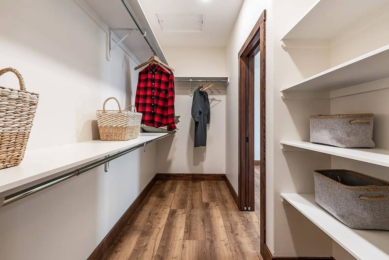 A spacious walk-in closet features wooden flooring, white shelves, and rails with a red plaid shirt and gray jacket. Baskets add an organized feel.
