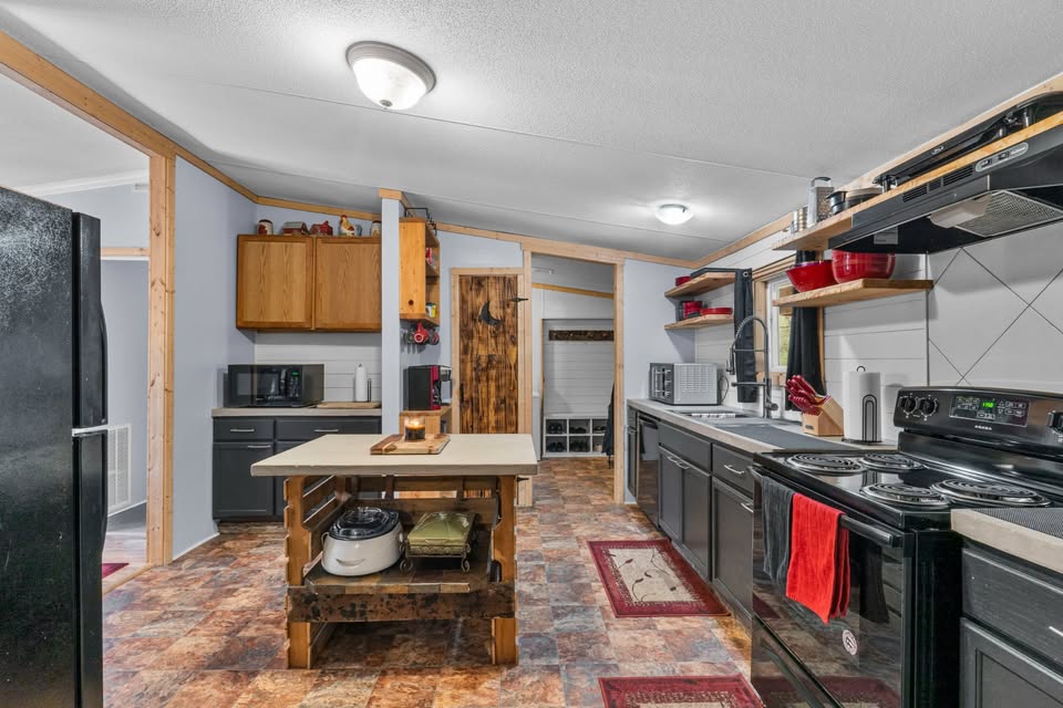 Rustic kitchen with black appliances, wooden island, and tiled floor. Open shelving holds red accents; cozy, functional vibe. Bright overhead lighting.
