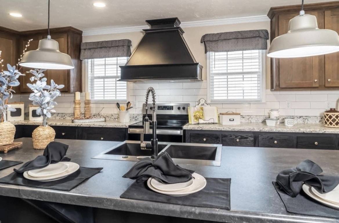 Modern kitchen interior with a black central island featuring place settings with black napkins, pendant lights, stainless steel appliances, and dark wooden cabinets.