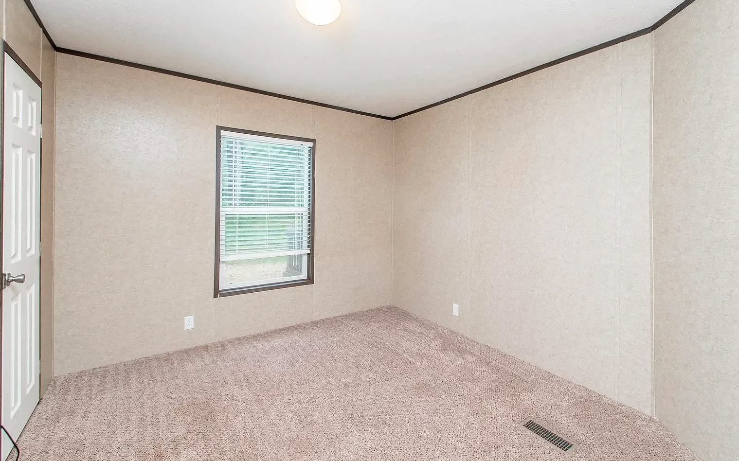 An empty, beige carpeted room with light brown walls, a single window with blinds, a door on the left, and a ceiling light, conveying a neutral tone.