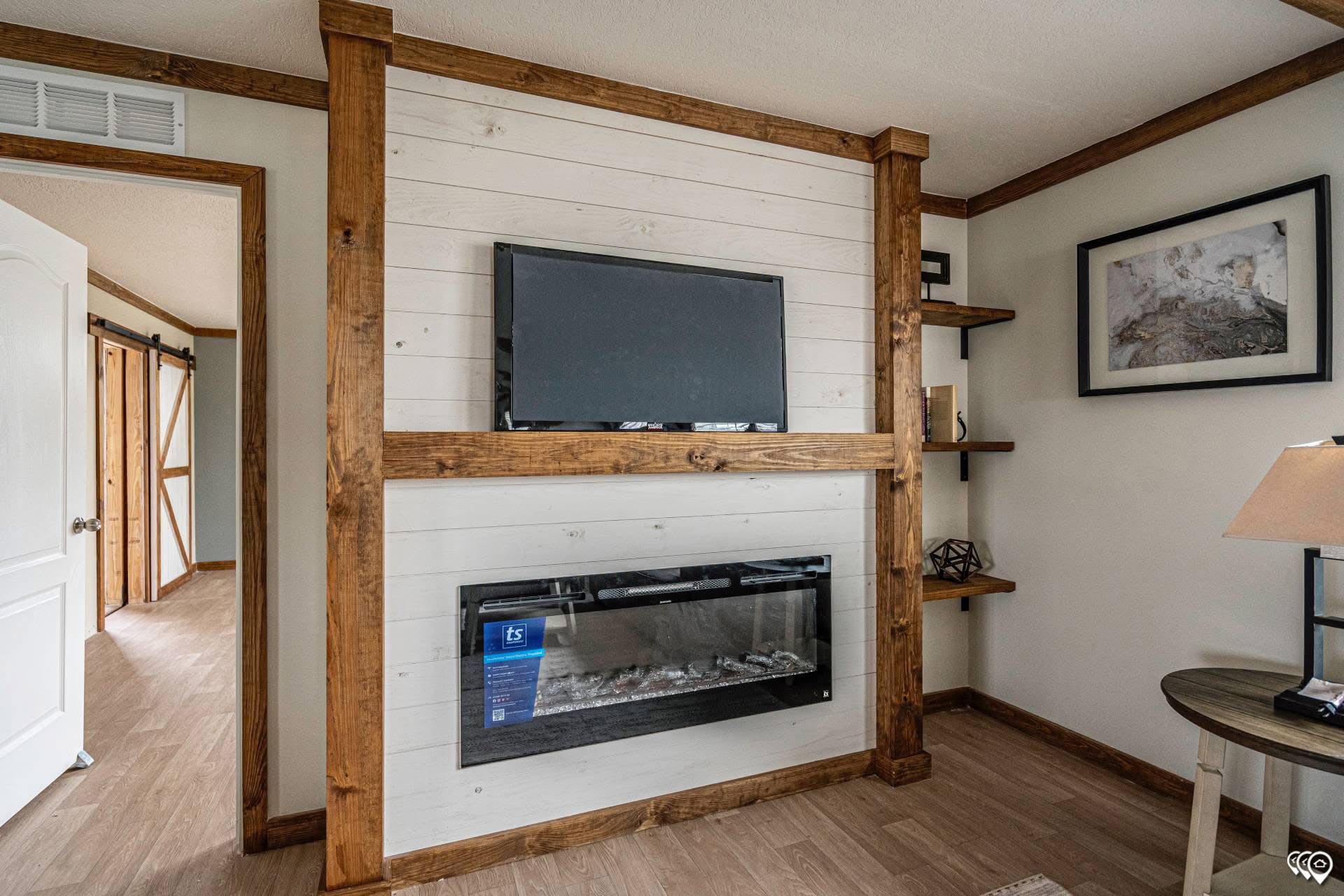 Modern living room with a rustic touch, featuring a wall-mounted TV and electric fireplace. Wooden shelves hold decor. Bright, inviting atmosphere.