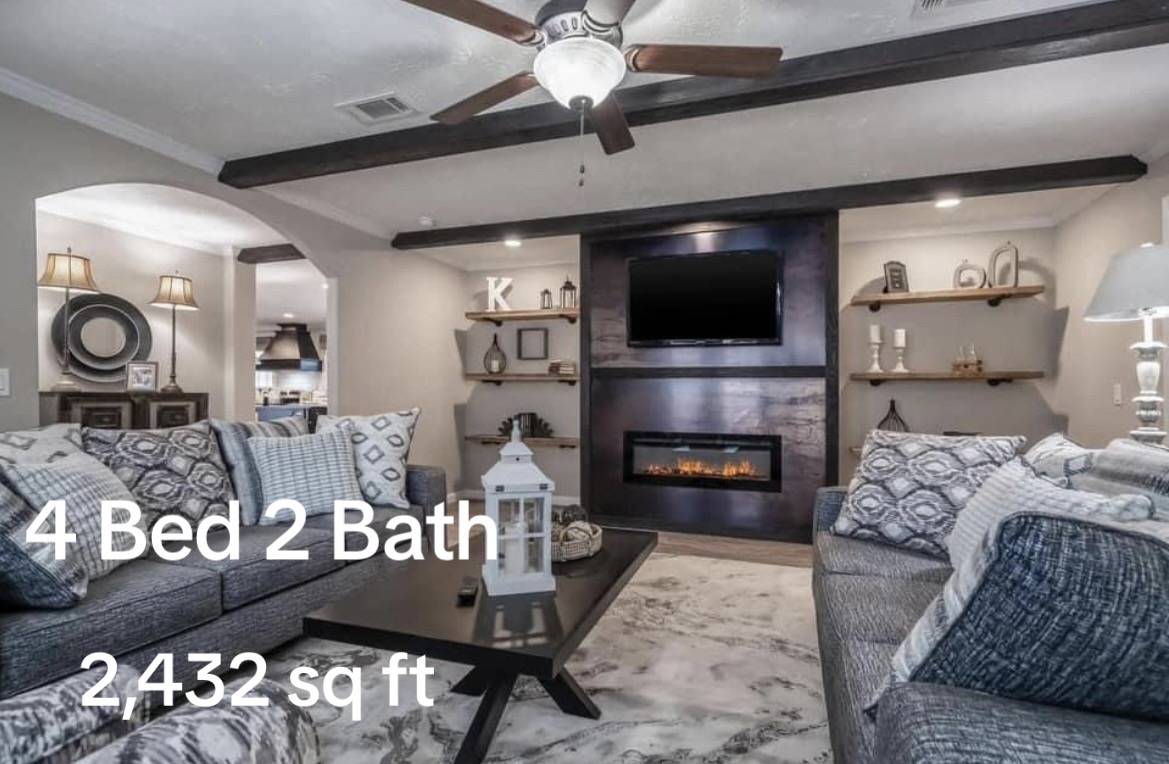Cozy living room with gray sofas, a large ceiling fan, and a fireplace under a wall-mounted TV. Warm lighting and neutral tones create a welcoming atmosphere. Text overlay: "4 Bed 2 Bath, 2,432 sq ft."