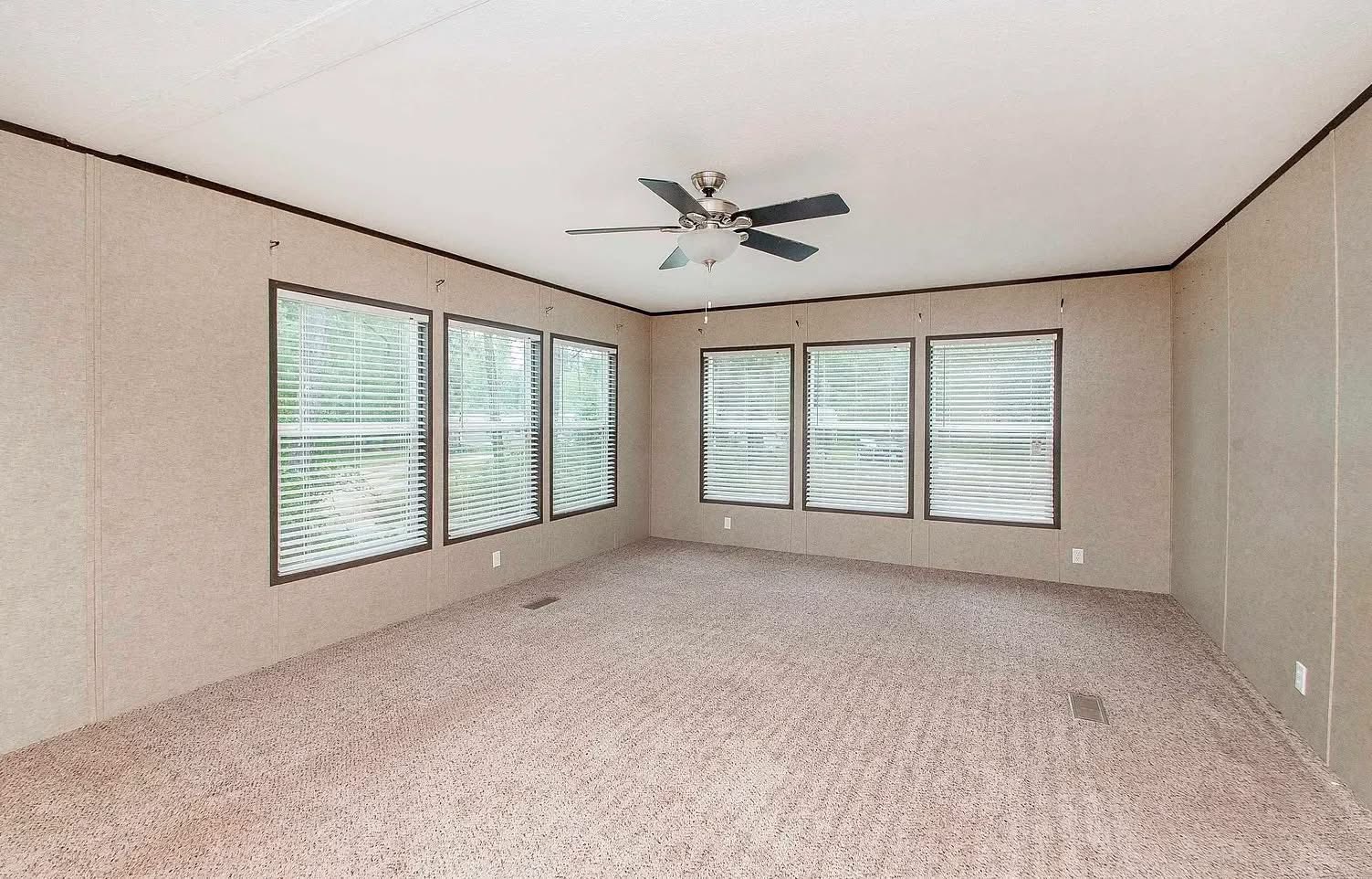 Spacious empty room with beige textured walls and carpet, featuring five large windows with blinds and a central ceiling fan, creating a calm and airy atmosphere.