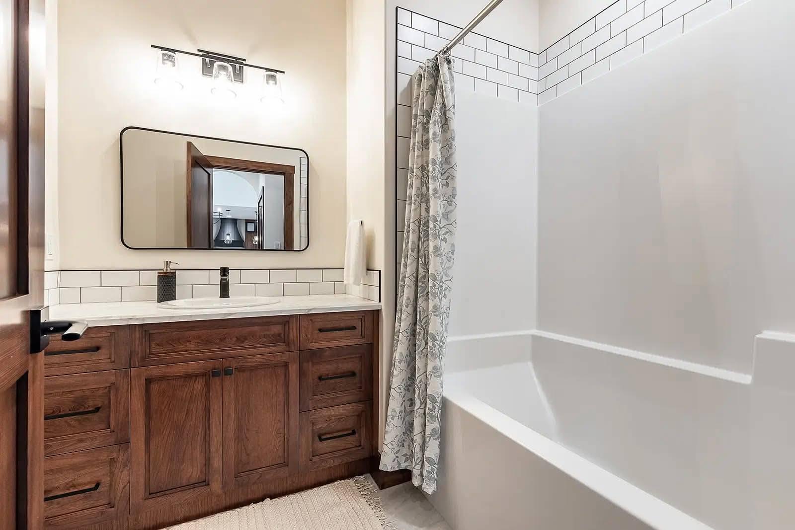A bathroom features a wooden vanity with a rectangular mirror and modern lighting. There is a white tiled wall, a bathtub with a floral shower curtain, and a cozy, clean aesthetic.
