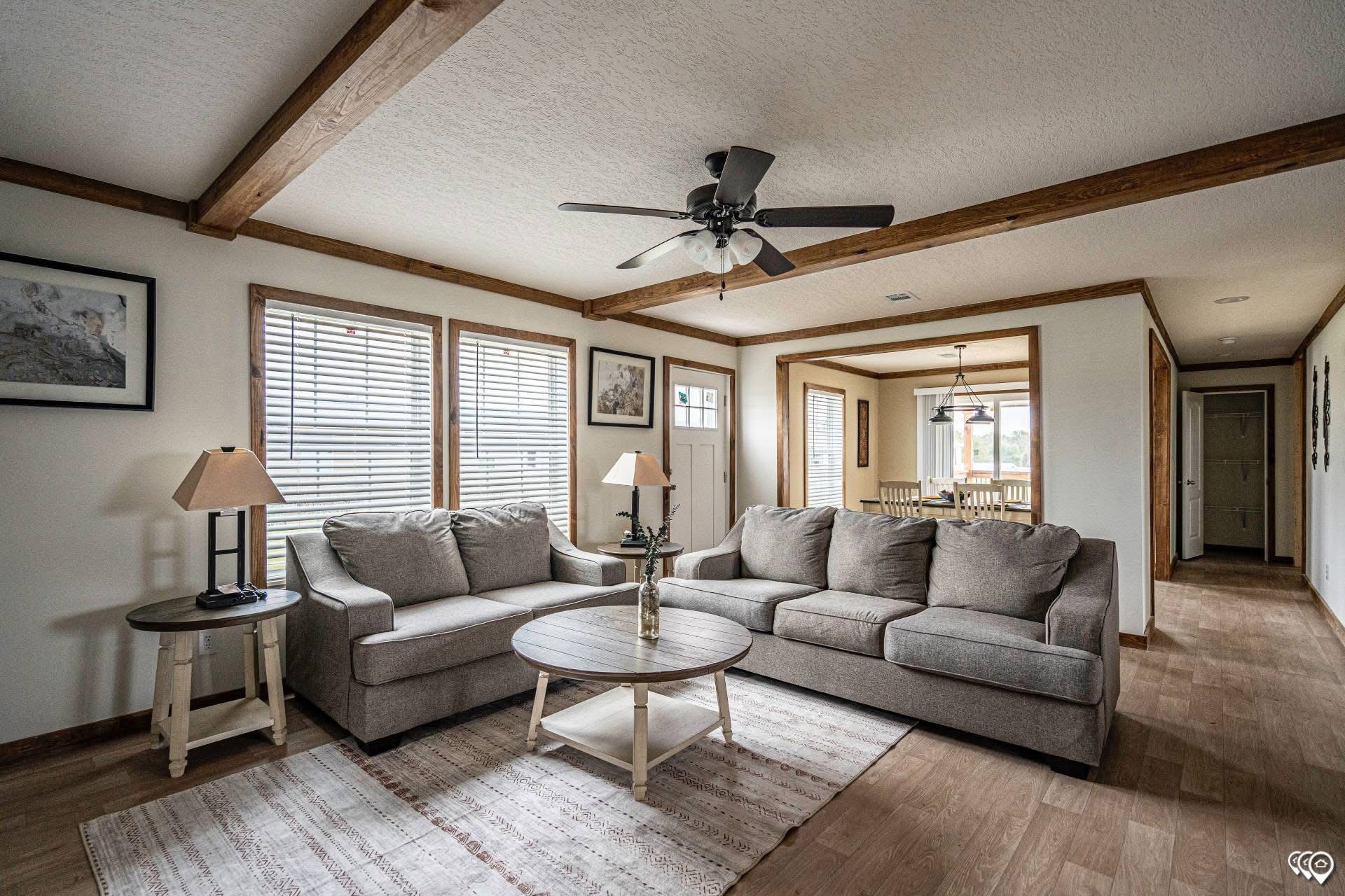 Bright living room with grey sectional sofa, wooden beams, ceiling fan, and framed art. Warm, inviting tone with a view into dining area.