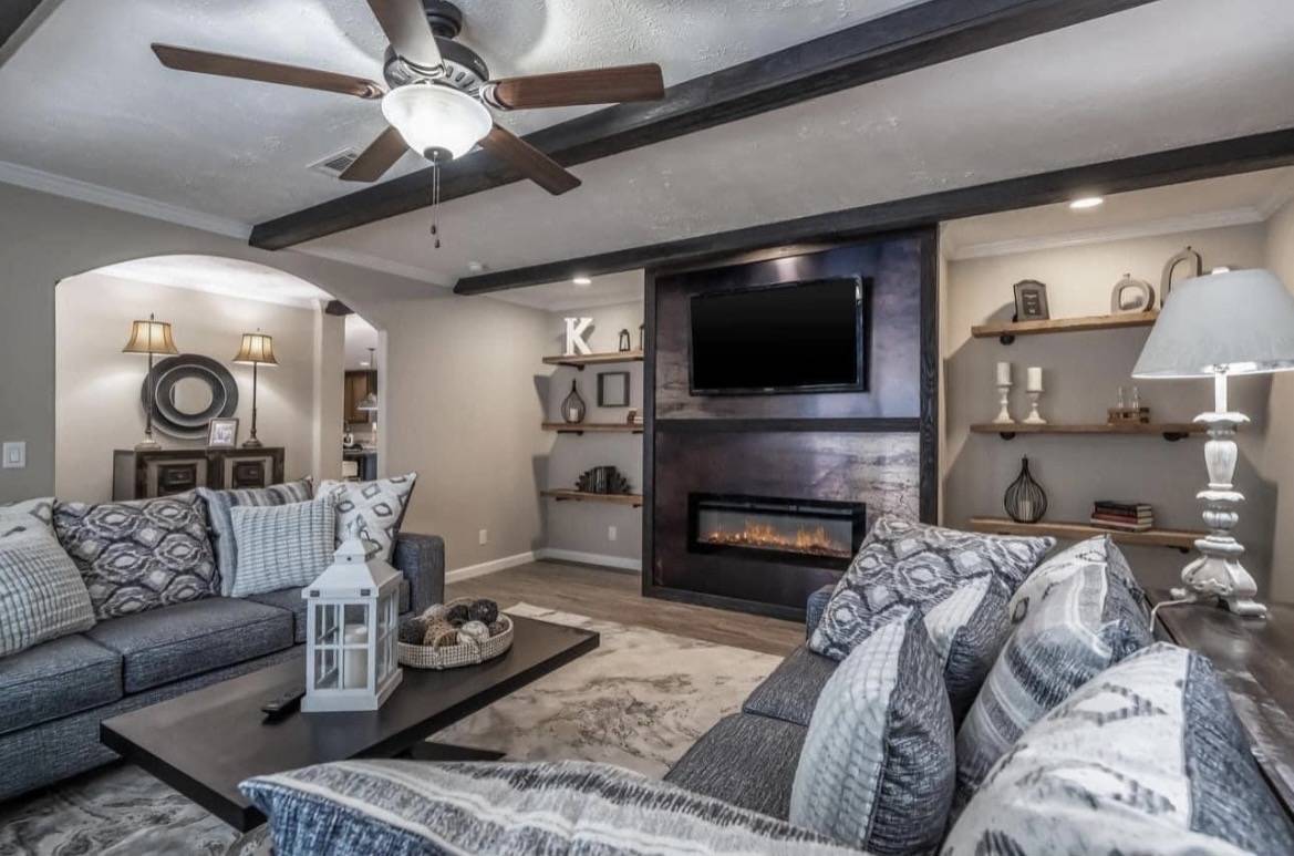 Cozy living room with a ceiling fan, gray couches adorned with patterned cushions, a wall-mounted TV, and an electric fireplace. Warm, inviting ambiance.