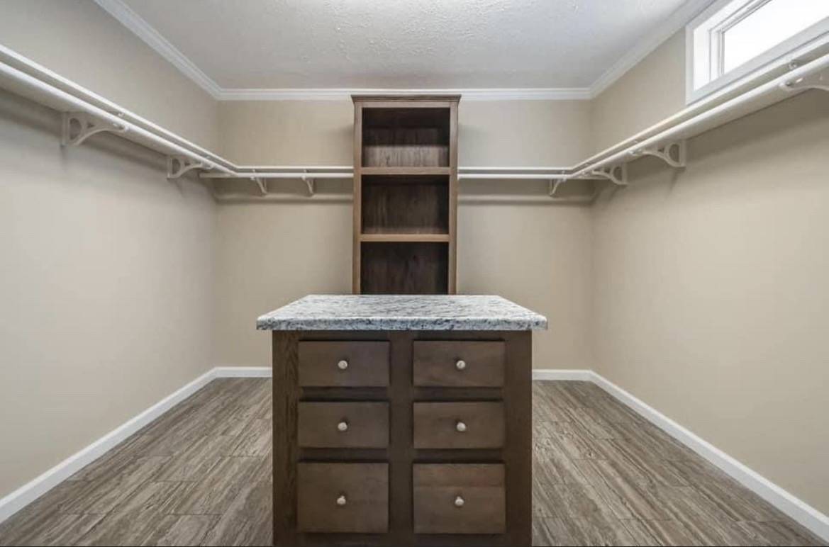 Spacious walk-in closet with beige walls, wooden floors, and overhead shelving. A wooden island with granite top and drawers is centered. Bright and orderly atmosphere.