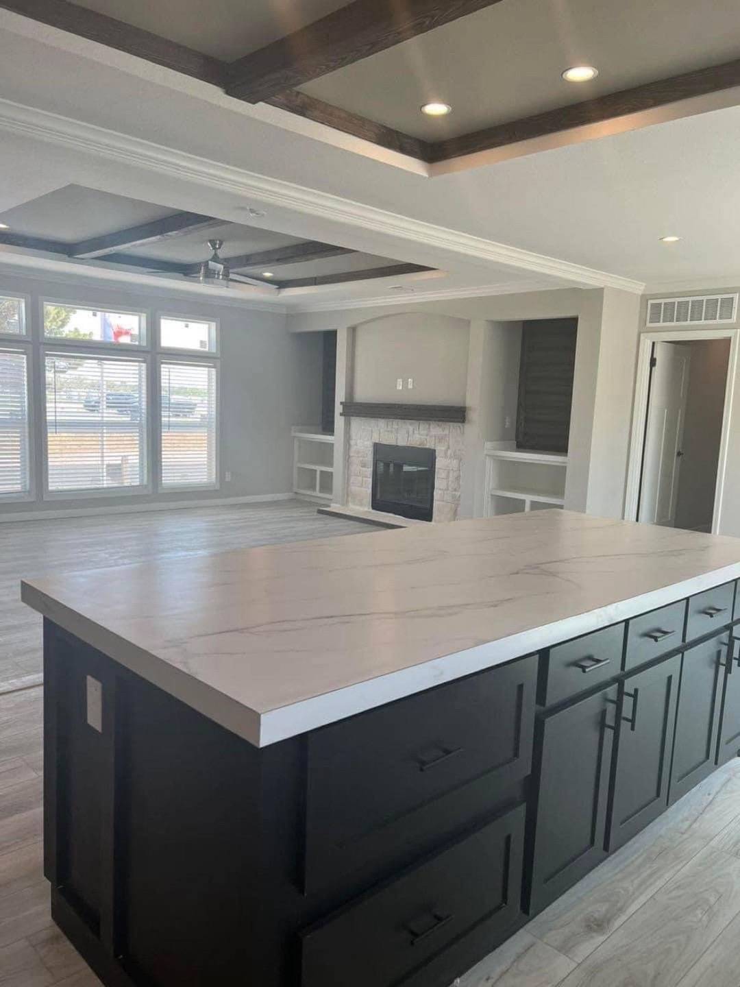 Spacious modern living room with a marble kitchen island in the foreground, wood-beamed ceiling, recessed lighting, large windows, and a fireplace.