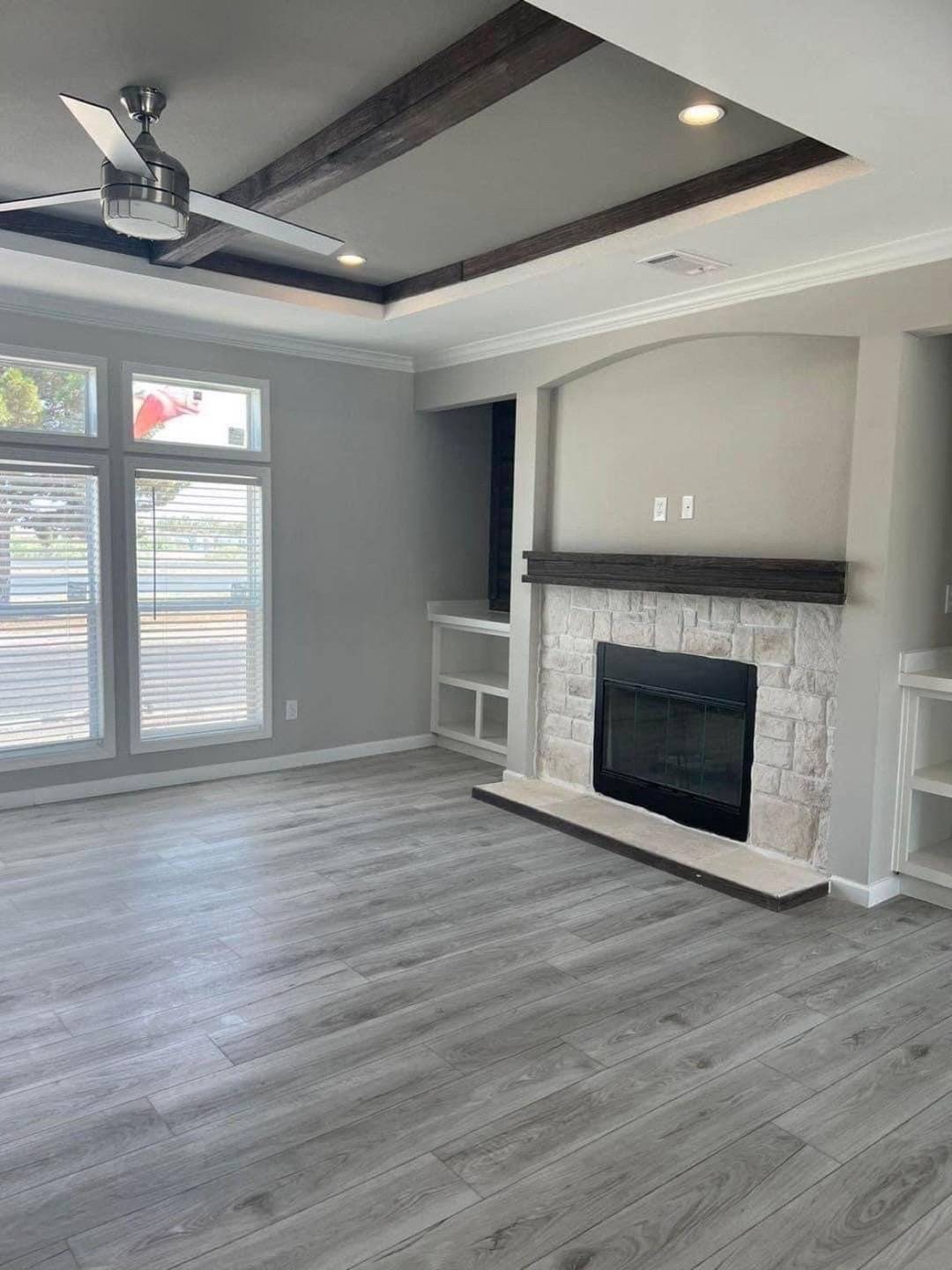 Modern living room with light wood flooring, stone fireplace, recessed shelves, and large windows. Ceiling features exposed beams and a fan.