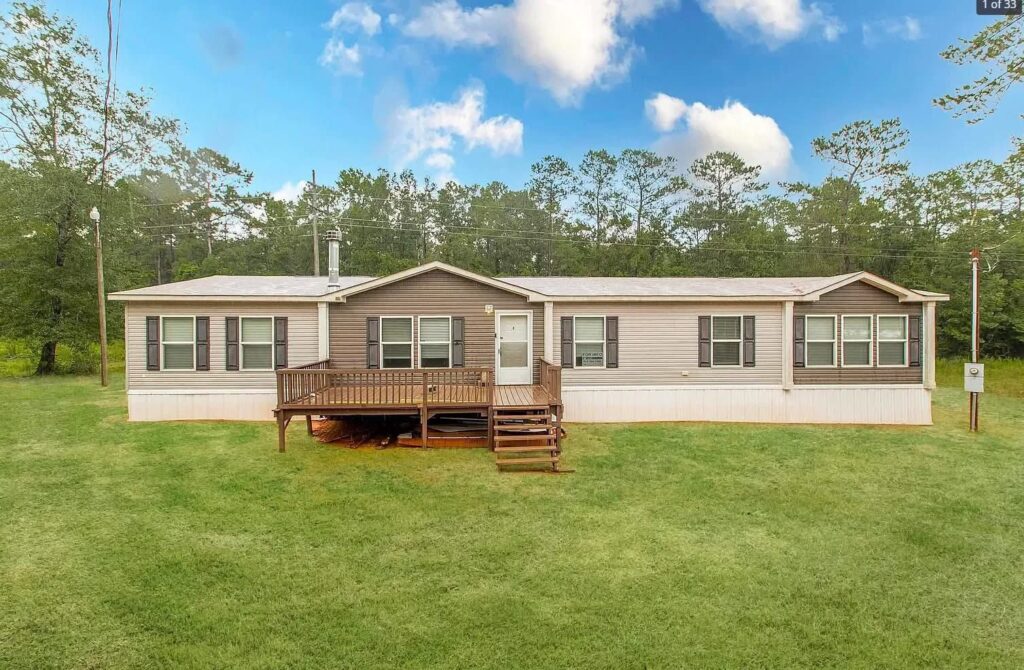 A beige mobile home with a wooden deck is set on a grassy lawn, surrounded by trees under a blue sky with fluffy clouds, conveying a peaceful, rural vibe.