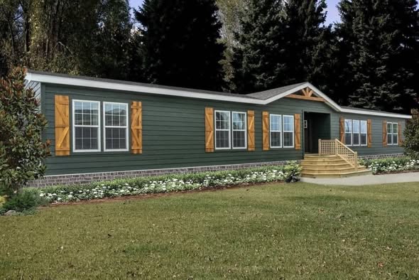 A long, dark green modular home with white-framed windows and wooden shutters. It has a small porch, surrounded by lush grass and tall trees.