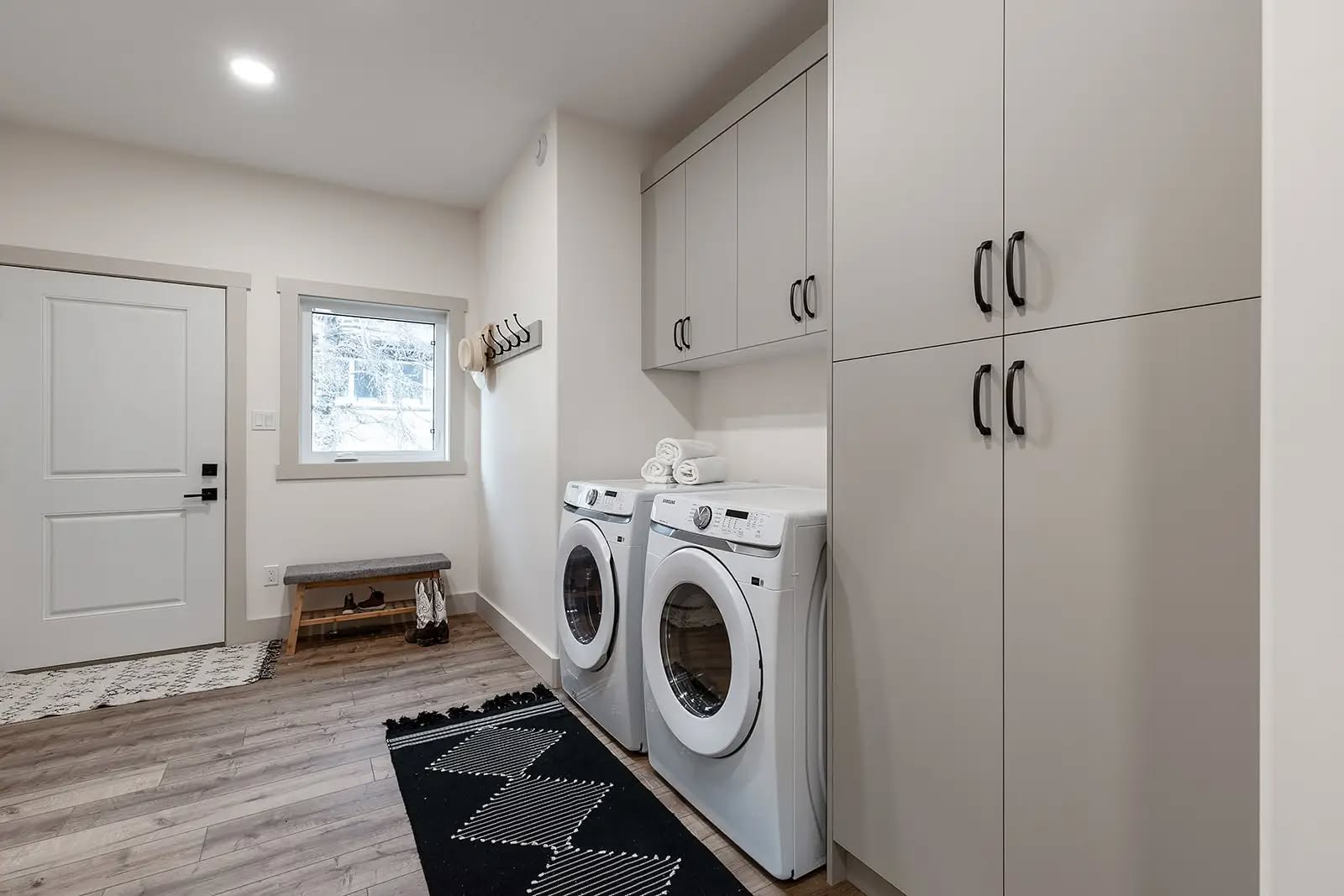 Modern laundry room with white walls, wooden floor, and recessed lighting. Features a washer and dryer, cabinets, and black rug. Bright and organized.