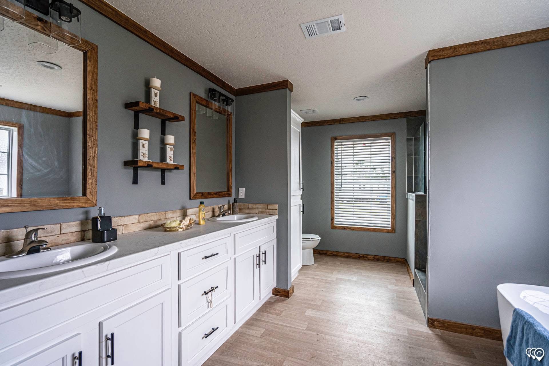 Modern bathroom with light wood flooring, double white vanity with sinks, two mirrors framed in wood, and shelves with candles. Calm and minimalist ambiance.
