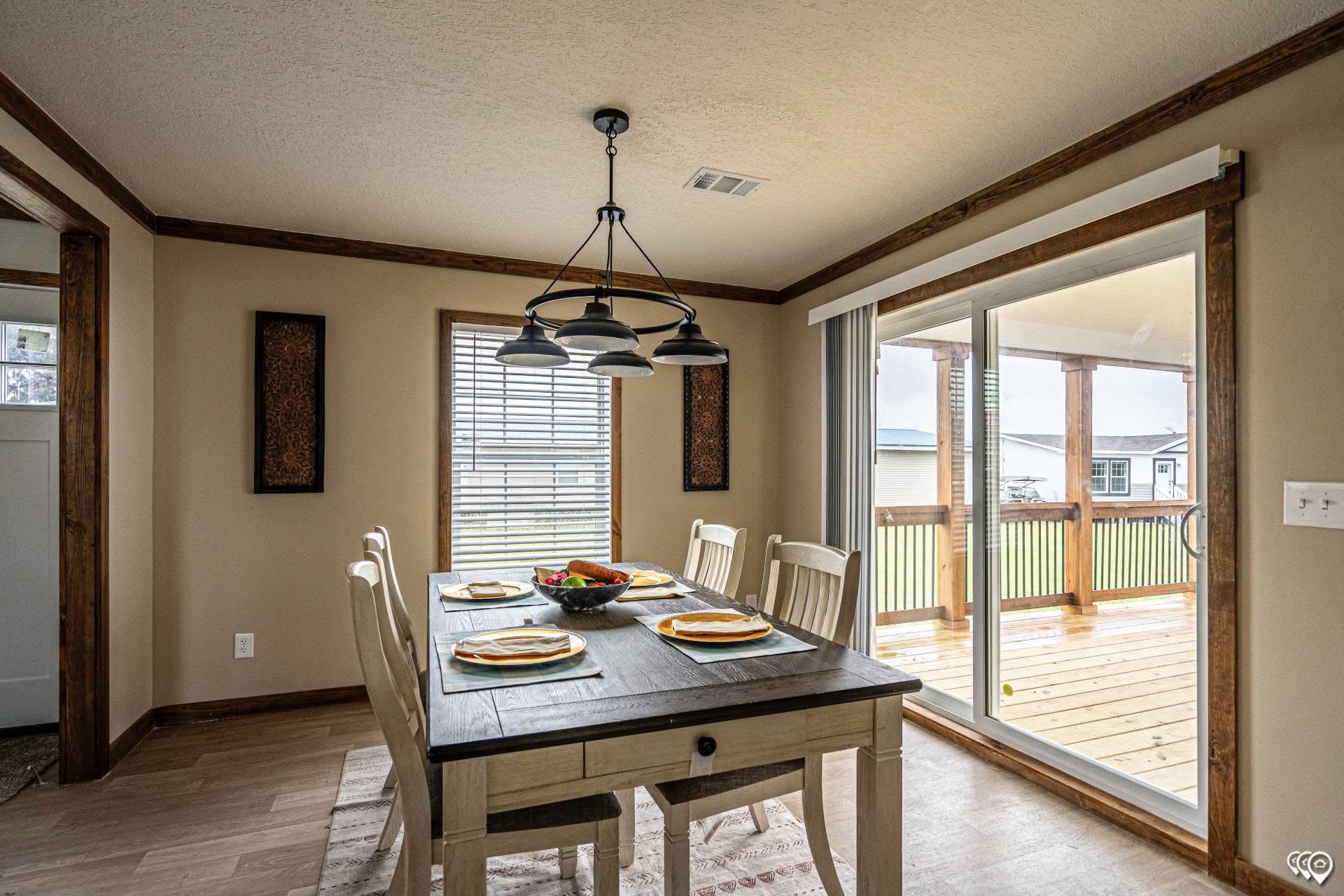 A cozy dining room with a rustic table set for four, featuring a fruit bowl centerpiece. Natural light streams through a large sliding glass door.