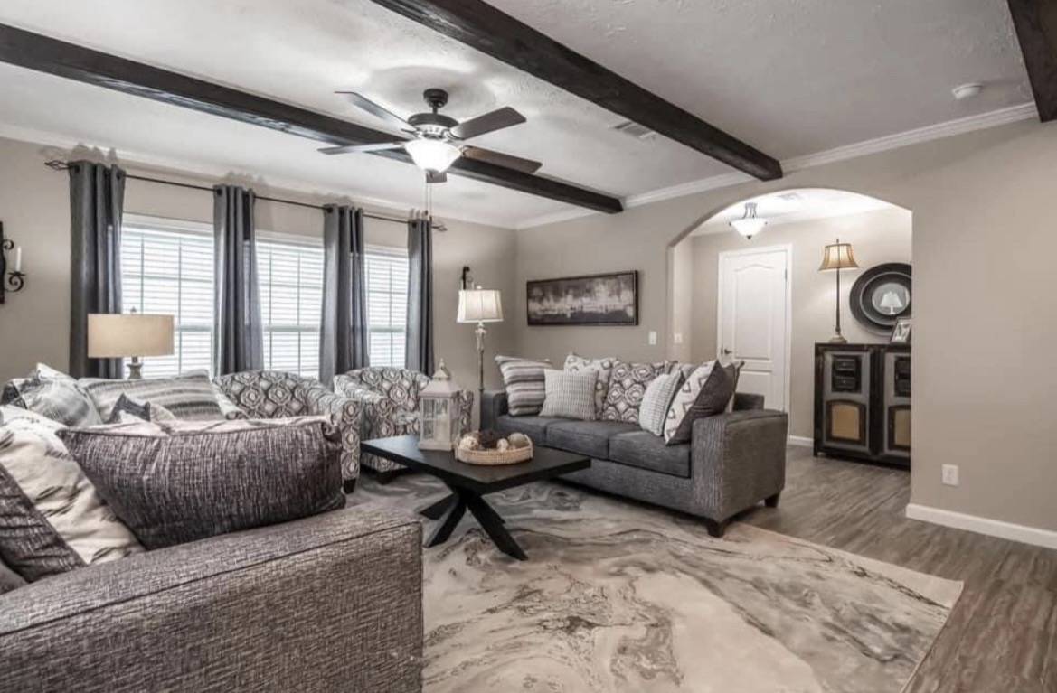 Stylish living room with gray couches, large windows, wooden beams on ceiling, and a patterned rug. Warm lighting creates a cozy, inviting atmosphere.