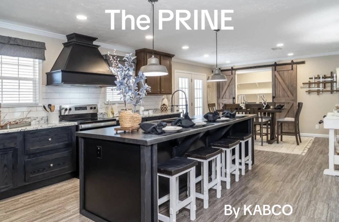 Modern kitchen with a black island, white stools, and stainless steel accents. Warm lighting and rustic barn doors add a cozy feel. Text: "The PRINE By KABCO".