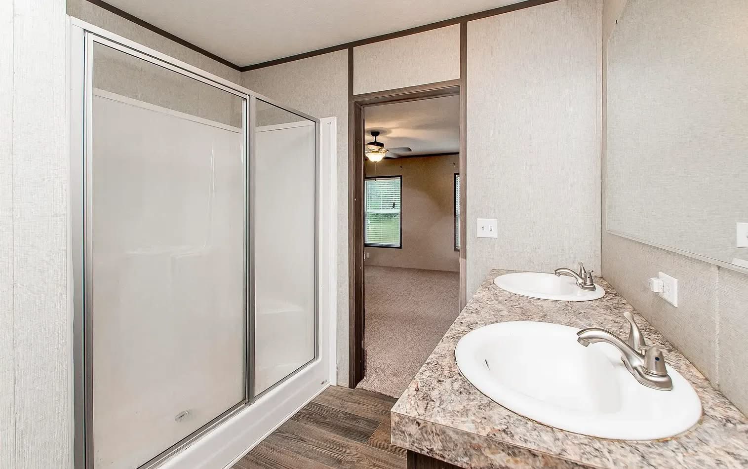 Modern bathroom with a dual-sink vanity on the right, spacious walk-in shower on the left, and an open doorway leading to a carpeted room with a ceiling fan.