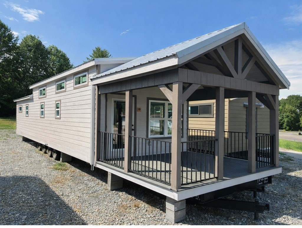 A modern tiny house with a gabled roof and large porch, set on gravel with green trees in the background under a clear blue sky.