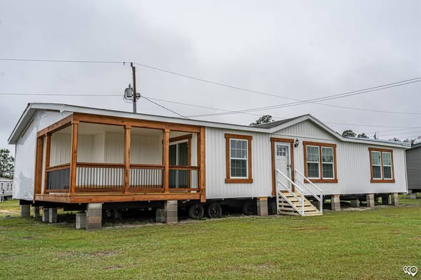 Single-story mobile home with white siding, large windows, and a covered wooden porch. It rests on cinder blocks in an open grassy area under a cloudy sky.