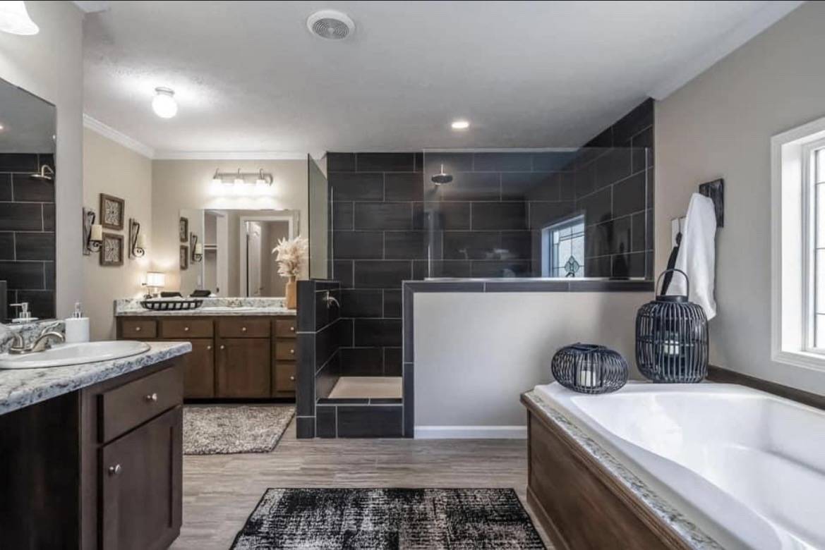 Spacious modern bathroom with a large bathtub, dark tile walk-in shower, double vanity with marble countertop, wooden cabinets, and soft lighting.
