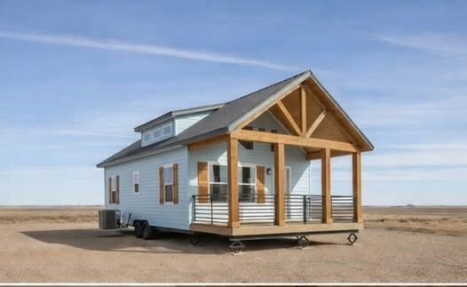 A light blue tiny house with a gabled roof and a wooden porch sits isolated in a vast, barren landscape under a clear blue sky. It exudes simplicity and tranquility.