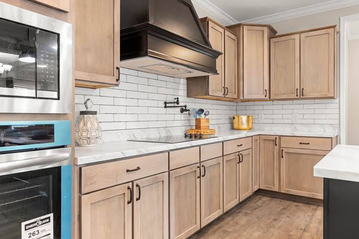 A modern kitchen with light wooden cabinets, white subway tile backsplash, and marble countertops. Stainless steel oven, dark range hood, and wooden floor add contrast. Accessories include a vase and utensils, creating a warm, functional space.