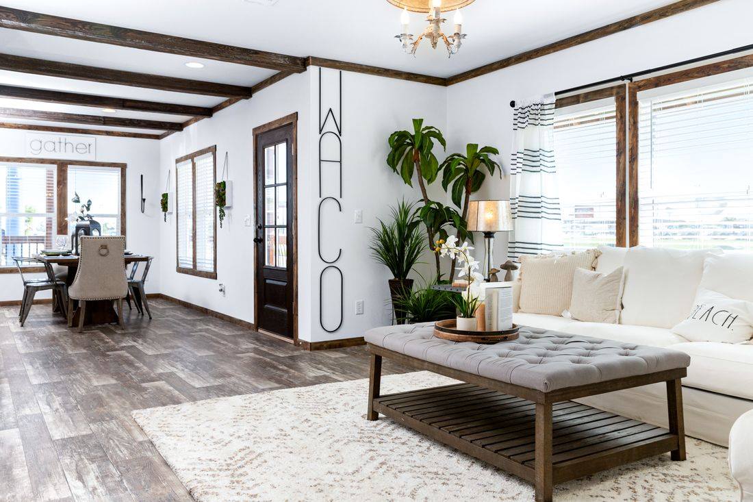 Spacious open-concept living and dining room in a luxurious modern farmhouse modular home featuring vaulted ceilings with exposed dark rustic wooden beams, crisp white shiplap walls accented by bold vertical black metal "WACO" sign and "gather" wall art, plush white sectional sofa layered with neutral textured pillows and beach-themed throws, tufted gray ottoman coffee table with wooden base and tray decor including potted orchids candles and books, large windows with sheer striped curtains flooding natural light, dark wood-look plank flooring, elegant chandelier and wall sconces, potted palm trees and greenery creating a warm inviting family gathering space perfect for relaxation entertaining and coastal-inspired living in this dream customizable manufactured home design.