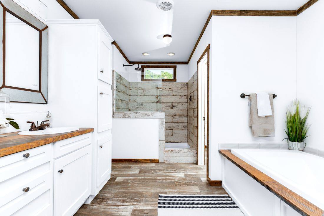 Spacious modern farmhouse master bathroom in a customizable modular tiny home featuring bright white shaker cabinets with oil-rubbed bronze hardware, rich reclaimed wood-look butcher block countertops on double vanity and soaking tub surround, elegant undermount oval sink with vintage-style faucets, large frameless mirror with rustic wood frame, walk-in frameless glass shower with distressed shiplap tiled walls seamless rainfall showerhead and step-up tiled bench, freestanding white oval bathtub positioned for natural light from small window, vaulted ceiling with exposed dark wooden beams recessed lighting and exhaust fan, warm wood-look plank flooring accented by striped black-and-white runner rug, potted greenery and plush white towels creating a serene spa-like retreat perfect for relaxation and rejuvenation in this dream affordable manufactured home design.