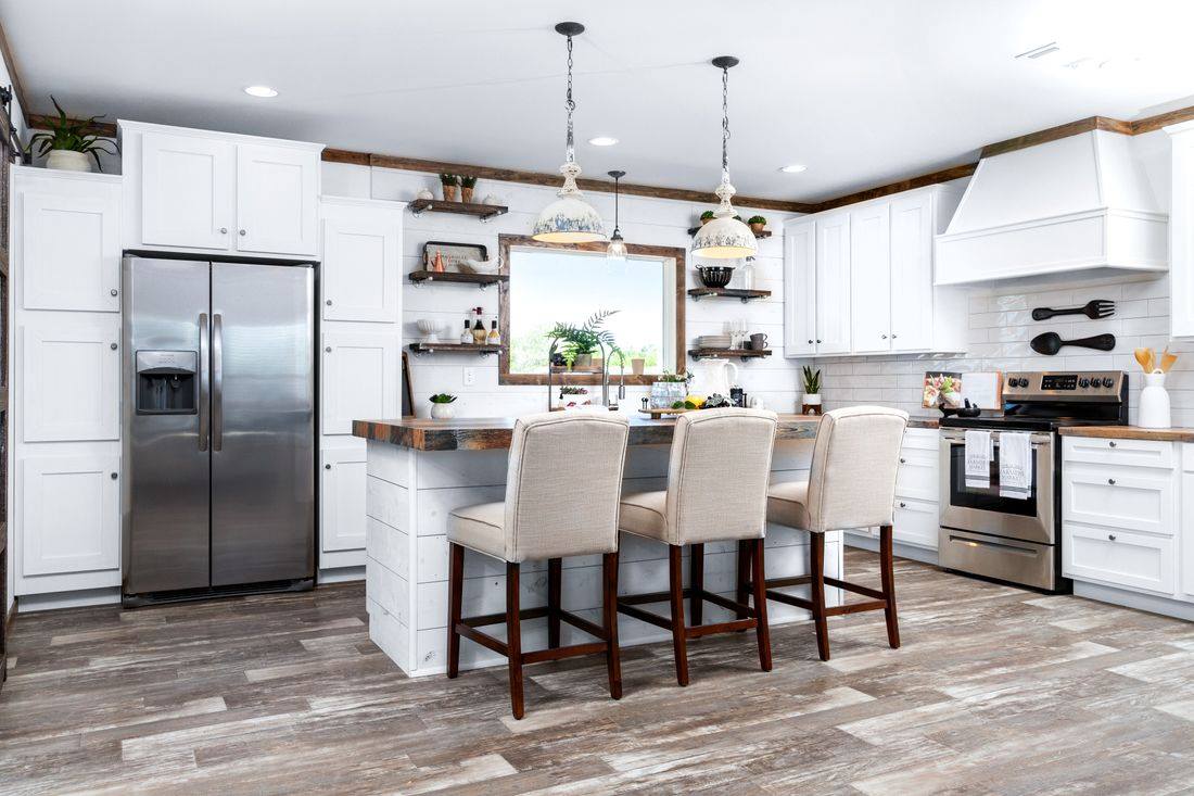 Luxurious open-concept farmhouse kitchen in a spacious customizable modular home featuring bright white shaker cabinets with black hardware, stunning reclaimed wood-look butcher block island countertop seating four elegant upholstered beige bar stools, large central island with deep sink farmhouse faucet and abundant prep space, stainless steel double-door refrigerator with water dispenser, gas range with black hood and subway tile backsplash, vaulted ceiling with exposed rustic wooden beams vintage pendant lights and modern ceiling fan, open shelving displaying potted herbs cookbooks and decor, large framed window with greenery views flooding natural light, warm wood-look plank flooring creating an inviting bright functional family cooking entertaining heart of the home in this dream affordable manufactured house design.