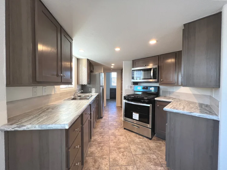 Spacious galley kitchen featuring dark wood cabinetry, granite-look countertops, stainless steel appliances including a gas range and microwave, double sink, and tiled flooring, with a view down a hallway towards another room.