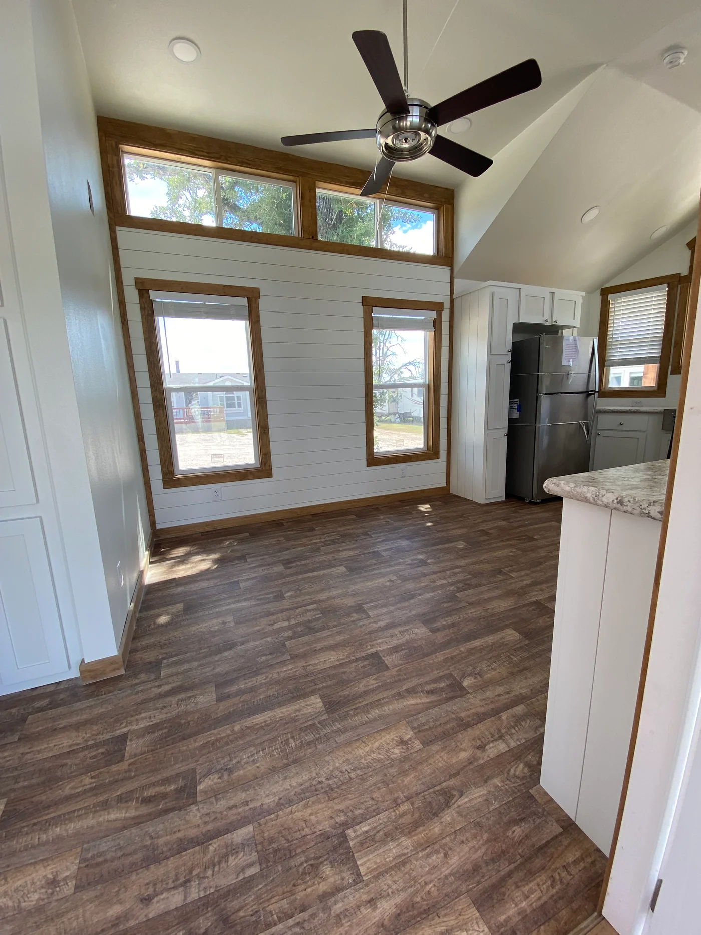 Modern luxury park model tiny home interior featuring an open-concept living space with high vaulted ceilings, white shiplap walls, and premium wide-plank wood flooring. The sophisticated architectural design includes energy-efficient windows with natural wood trim, a contemporary stainless steel ceiling fan, and a gourmet kitchen area equipped with a stainless steel refrigerator and elegant stone-patterned countertops. This bright and airy prefab unit captures a professional "full rank" SEO-optimized aesthetic, making it an ideal showcase for custom-built small-space living, high-end vacation cottage interiors, or modern backyard accessory dwelling units (ADU) with maximum natural light and a clean, minimalist feel.