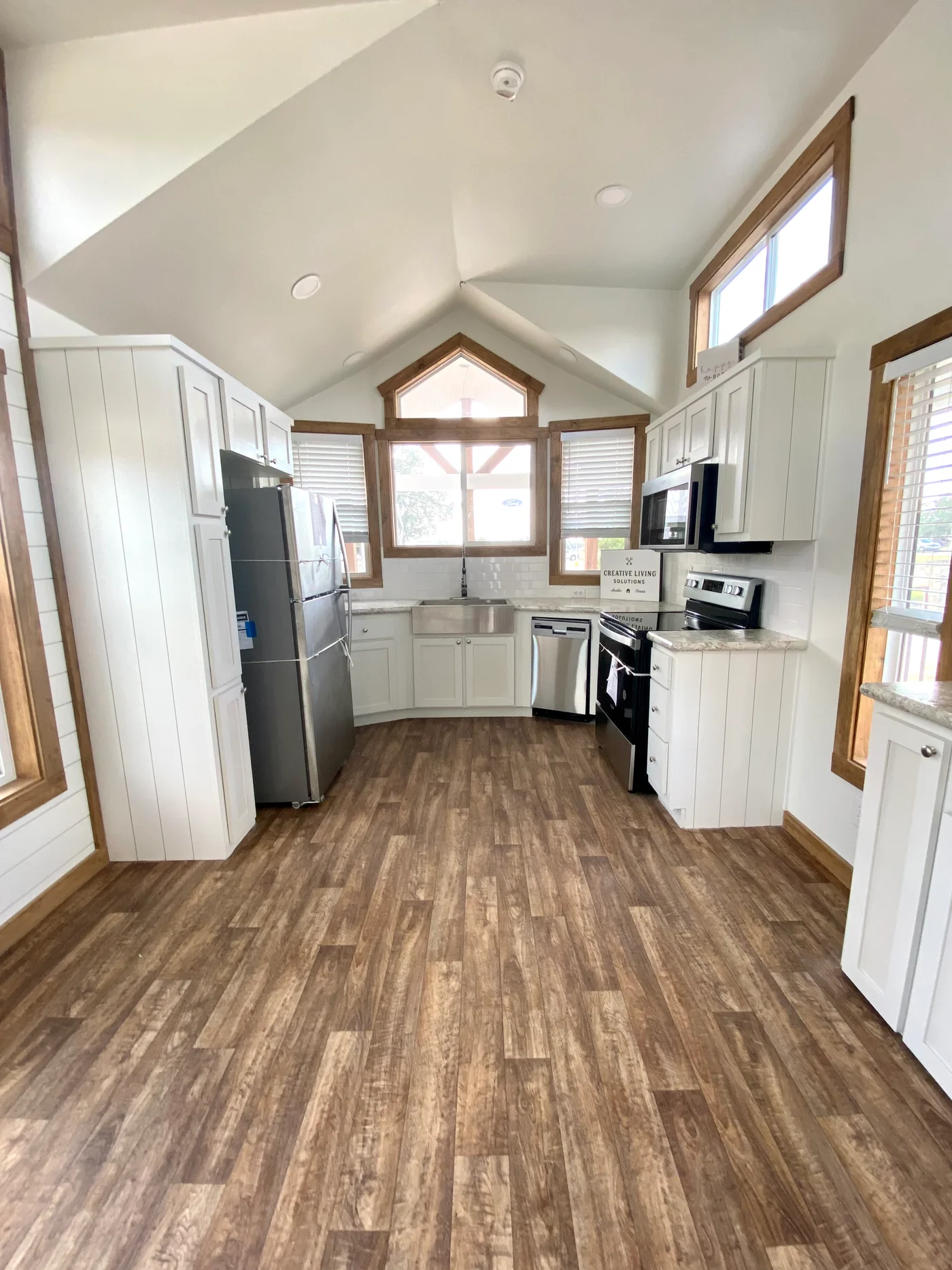 Modern luxury farmhouse-style tiny home kitchen featuring bright white shiplap walls, high vaulted ceilings, and premium wide-plank wood flooring that creates a spacious, airy feel. The sophisticated gourmet space is equipped with high-end stainless steel appliances, including a large refrigerator and dishwasher, complemented by elegant white cabinetry, a deep farmhouse-style sink, and a sleek black glass-top range. Large energy-efficient windows with natural wood trim flood the kitchen with natural light, highlighting the contemporary "full rank" SEO-optimized architectural design perfect for luxury prefab living, custom-built vacation cottages, or stylish accessory dwelling units (ADU) with a professional and clean aesthetic.