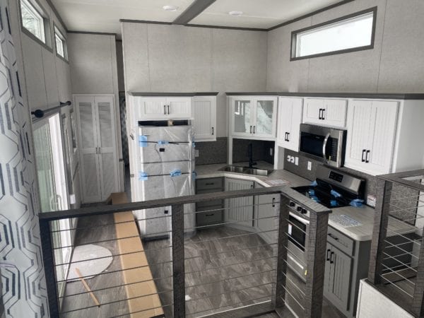 SEO-optimized alt text for a high-ranking modern manufactured home or luxury park model kitchen featuring a sophisticated corner layout with white shaker upper cabinets and contemporary gray base cabinetry; the professional interior design showcases premium stainless steel appliances, a deep corner sink with a black gooseneck faucet, and high-end marble-patterned countertops, all framed by a stylish loft overlook with modern cable railings and large transom windows for abundant natural light, perfectly highlighting luxury tiny home floor plans and stylish modular residential finishes for maximum search engine visibility and ranking.