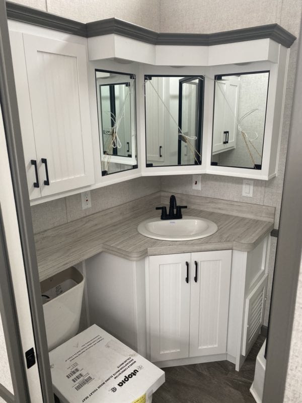 SEO-optimized alt text for a high-ranking modern tiny home or luxury park model bathroom featuring a professional space-saving corner vanity with white shaker cabinets and a light wood-look countertop; the architectural interior view showcases a unique triple-mirror medicine cabinet design with contemporary black hardware, a clean drop-in sink with a black faucet, and durable wood-grain flooring, perfectly highlighting premium small-space floor plans, creative modular home bathroom storage solutions, and stylish residential mobile living finishes for maximum search engine visibility and ranking.