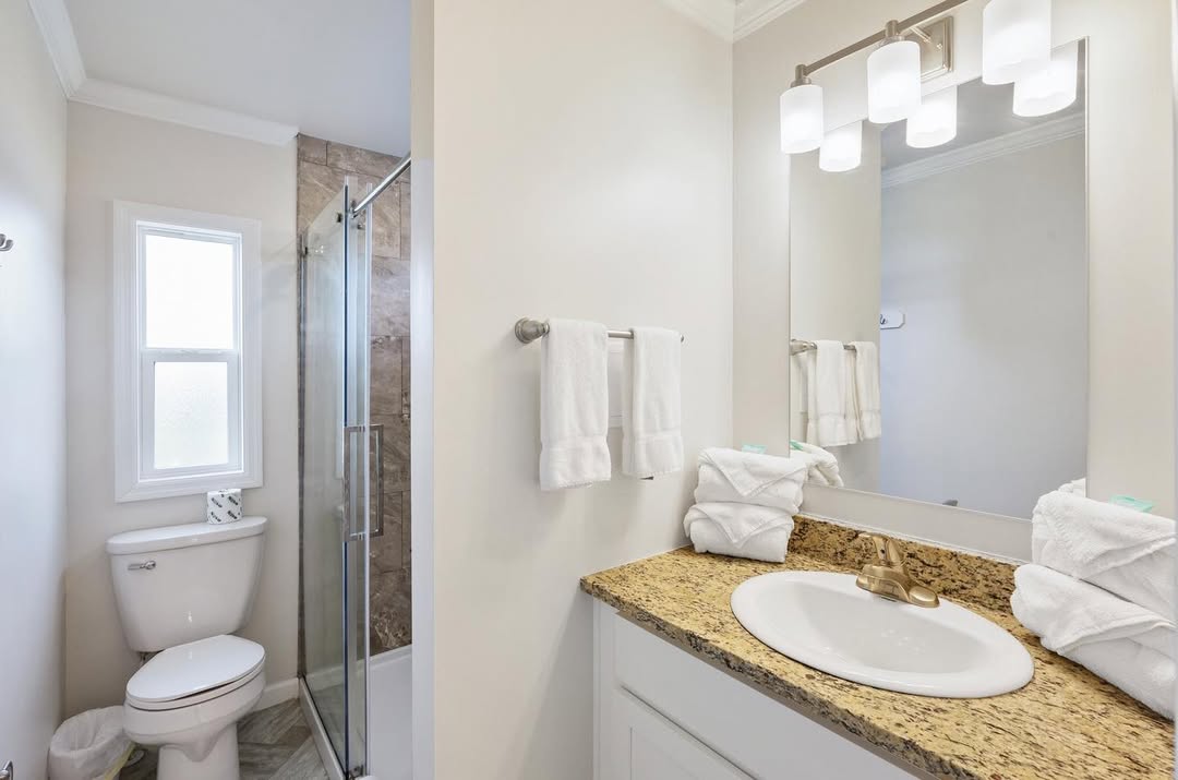 Bright and spacious modern bathroom in a customizable modular home featuring neutral beige walls with crisp white trim, elegant white shaker vanity cabinet with gold hardware and speckled granite countertop, undermount oval sink with brushed gold faucet, large rectangular mirror framed in white with matching gold-framed vanity lights providing warm illumination, frameless glass walk-in shower enclosure with seamless tiled walls in earthy tones, standard white toilet, small window allowing natural light and ventilation, plush white towels neatly folded on chrome towel bar, clean tiled flooring creating a fresh inviting functional guest or secondary bath retreat perfect for everyday use and relaxation in this affordable dream manufactured home design.