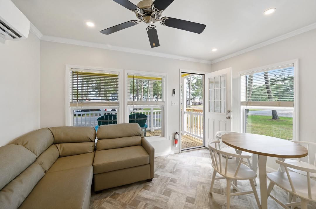 Cozy open-concept living and dining area in a bright modern tiny modular home featuring plush tan sectional sofa with recliner ends arranged for comfortable lounging, round light wood dining table with white spindle chairs seating four, vaulted ceiling with elegant black ceiling fan and recessed lighting, large wraparound windows and French doors with white blinds flooding the space with natural light and scenic outdoor views of deck trees and community, neutral beige walls white trim herringbone-pattern wood-look tile flooring seamless flow to exterior porch creating an inviting airy family-friendly gathering space perfect for relaxation meals and entertaining in this affordable compact dream manufactured tiny house design.