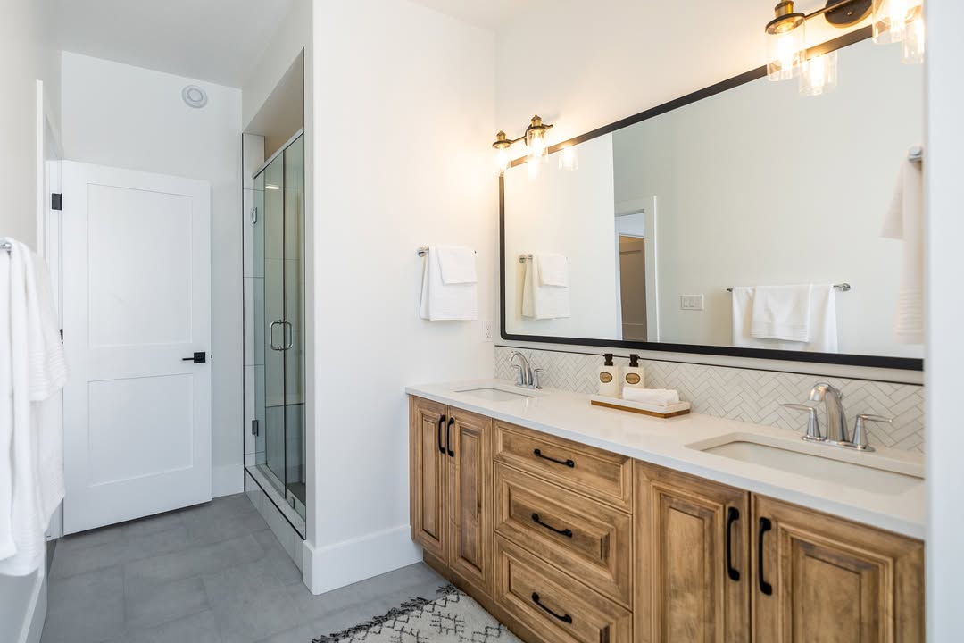 Modern luxury bathroom featuring a double vanity with natural wood cabinetry and black hardware, topped with a white quartz countertop and dual undermount sinks. The contemporary design includes a large black-framed mirror, elegant gold sconce lighting, and a white herringbone tile backsplash, complemented by a seamless glass walk-in shower and clean white walls for a bright, spa-like interior aesthetic.