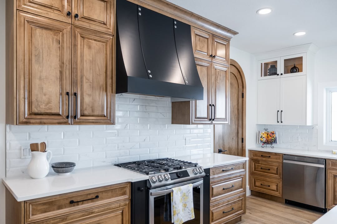 Modern luxury kitchen featuring custom warm wood cabinetry with black hardware, a sleek black designer range hood, and a white subway tile backsplash under bright recessed lighting. The contemporary interior design showcases high-end stainless steel appliances, white quartz countertops, and a glimpse into a butler's pantry through a unique arched wooden doorway, all set against light wide-plank hardwood flooring for a sophisticated farmhouse aesthetic.