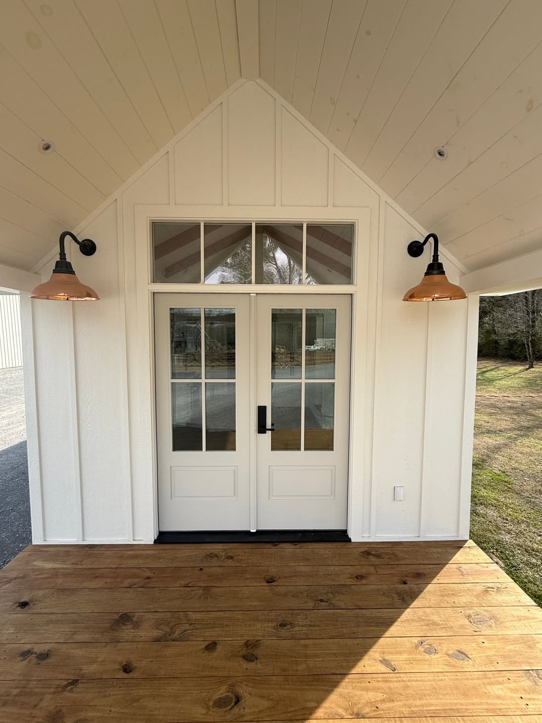Modern farmhouse entryway featuring elegant white double French doors with glass panes and a matching transom window, set beneath a high vaulted porch ceiling with white board and batten siding. The sophisticated architectural design is accented by two vintage-style copper gooseneck wall lanterns and a premium natural wood-plank deck, creating a bright and welcoming luxury entrance for a contemporary custom-built or high-end modular home.