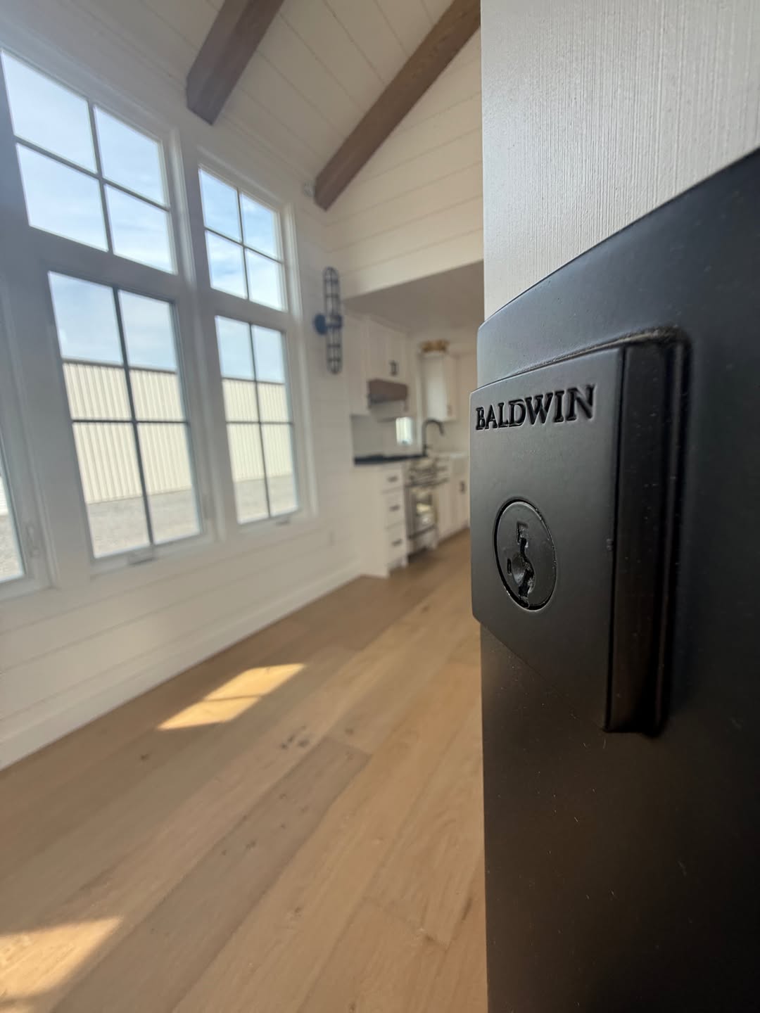A close-up shot of a high-end black Baldwin deadbolt door lock installed on a modern white door, showcasing luxury security hardware in a bright, open-concept home interior. The blurred background features premium light wood flooring, large white-trimmed windows, and a glimpse of a contemporary kitchen with a vaulted ceiling and rustic wood beams, highlighting the sophisticated architectural details and quality finishes of a custom-built or luxury modular farmhouse.