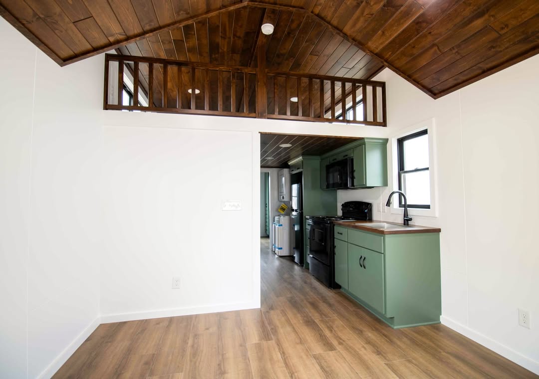 Compact kitchen in a tiny house with green cabinets, dark appliances, and wood flooring. Loft space above, featuring a wood ceiling and railing. Bright and cozy.