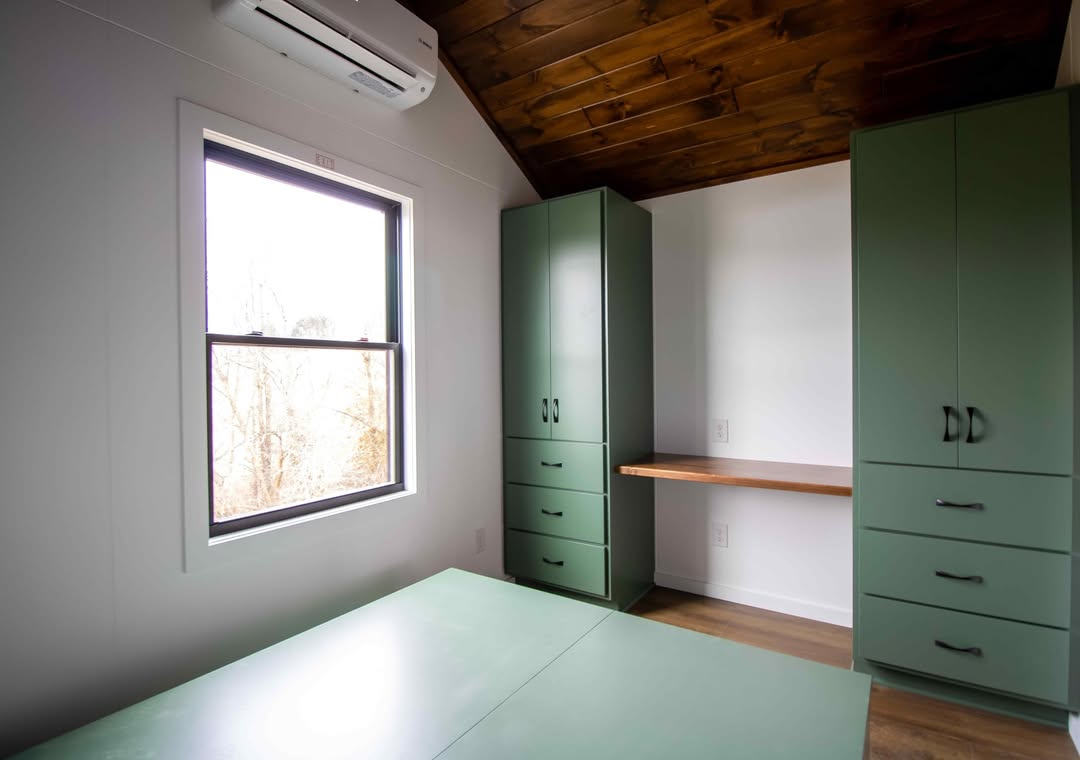Modern room with green cabinets, wooden ceiling, and large window. Bright light creates a fresh atmosphere. Minimalist desk under the window.