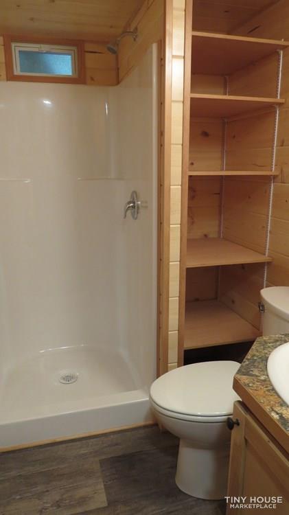 SEO optimized alt text: Tiny house bathroom with white corner shower, built-in shelf, metal showerhead, adjacent toilet, rustic wooden shelving unit, granite-style countertop, white sink basin, light wood walls, vinyl wood-look flooring, and frosted window for natural light.