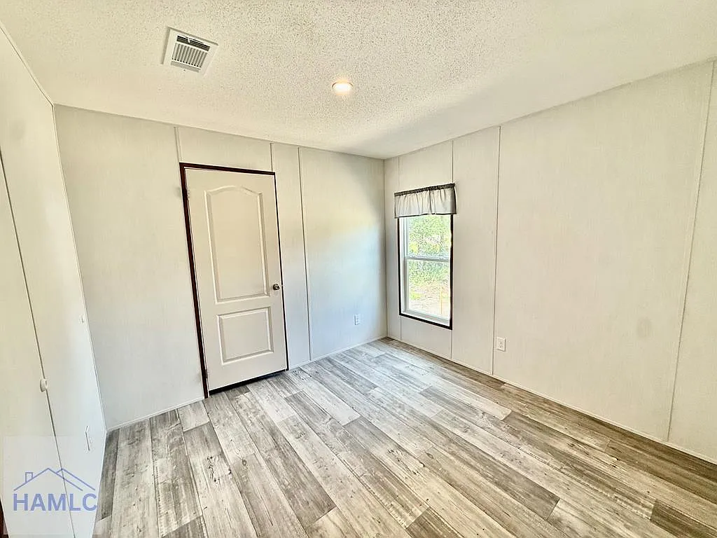 Spacious empty secondary bedroom in modern manufactured home featuring light neutral paneled walls with white trim, gray wood-look luxury vinyl plank flooring, textured popcorn ceiling with recessed lighting and HVAC vent, white arched interior door with dark trim, single large window with valance curtain overlooking green trees bringing natural light, bright airy unfurnished guest or kids room layout ready for personalization in premium 2025 double-wide modular home by HAMLC