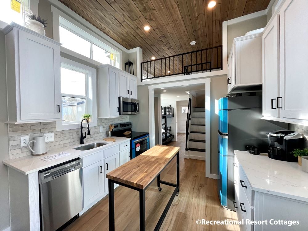 
Gemini said
Modern tiny house interior featuring a bright open-concept kitchen with white shaker cabinets, white quartz countertops, and a rustic wood-plank ceiling with recessed lighting. The space is optimized for small-scale living with a mobile butcher block kitchen island, a vibrant blue refrigerator, stainless steel appliances, and a loft sleeping area with black metal railings accessible by a staircase, showcasing a contemporary park model cottage design by Recreational Resort Cottages.