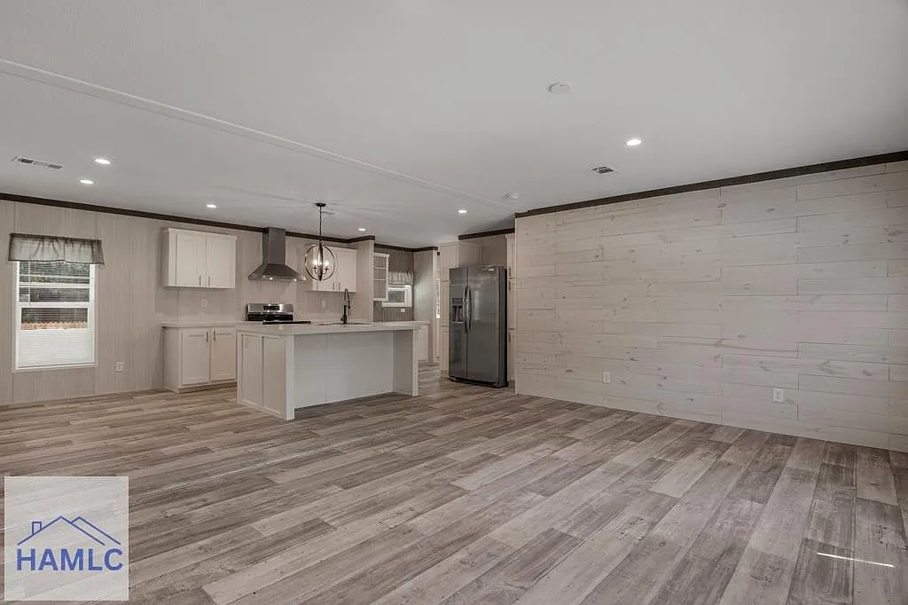 Spacious open-concept kitchen and living area in luxury modern manufactured home, vaulted ceiling with recessed lighting and tray detail, light beige shiplap accent walls with dark wood trim, large white shaker kitchen cabinets, expansive white quartz island countertop, stainless steel range hood and appliances including French door refrigerator, matte black farmhouse sink faucet pendant chandelier, gray wood-look luxury vinyl plank flooring throughout, bright natural light from windows with valance curtains, unfurnished airy great room layout in premium 2025 double-wide modular home by HAMLC